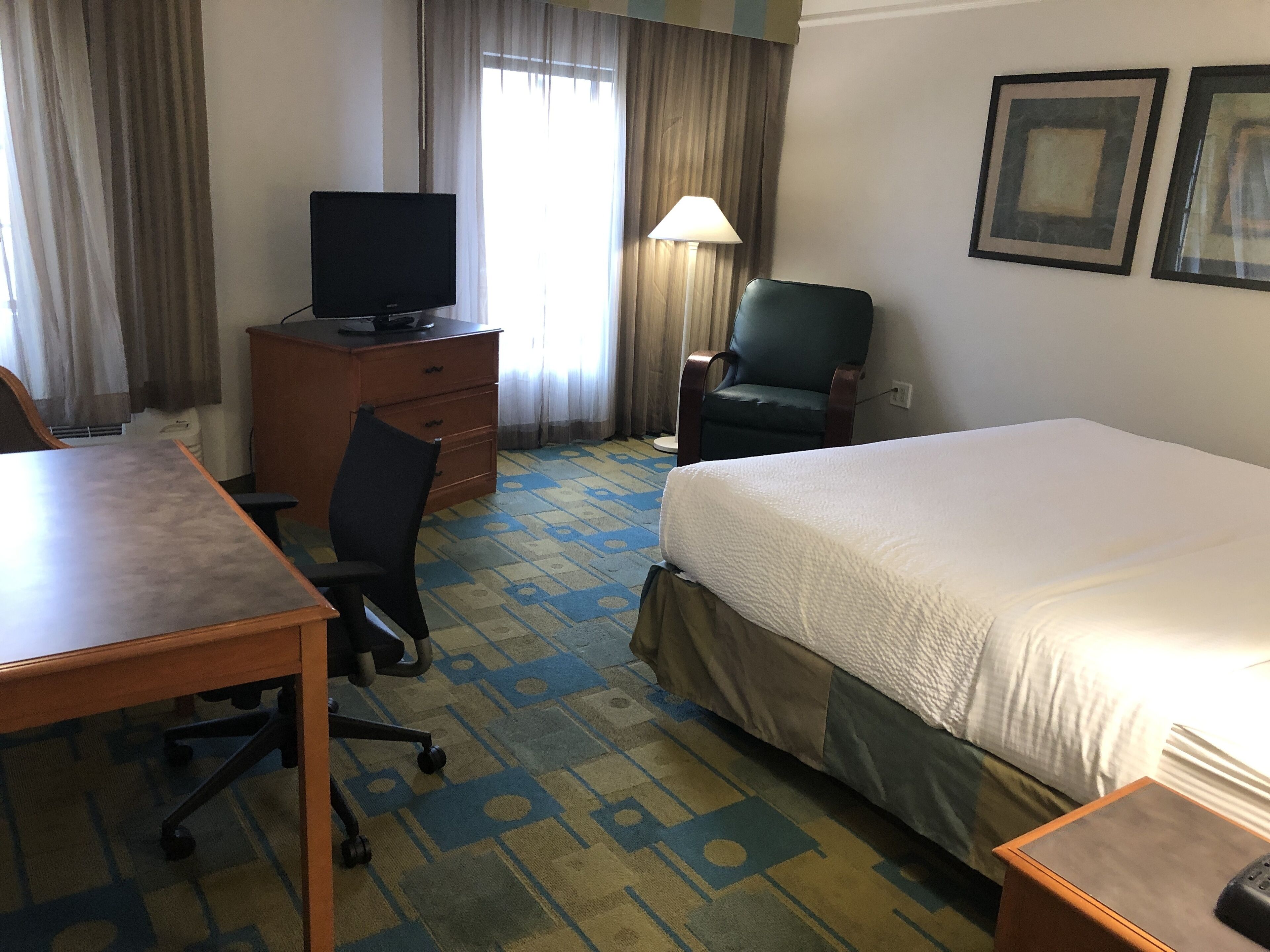 Een hotelkamer met een bed, een bureau, een stoel, een televisie op een dressoir, een lamp en twee foto's in lijsten aan de muur.