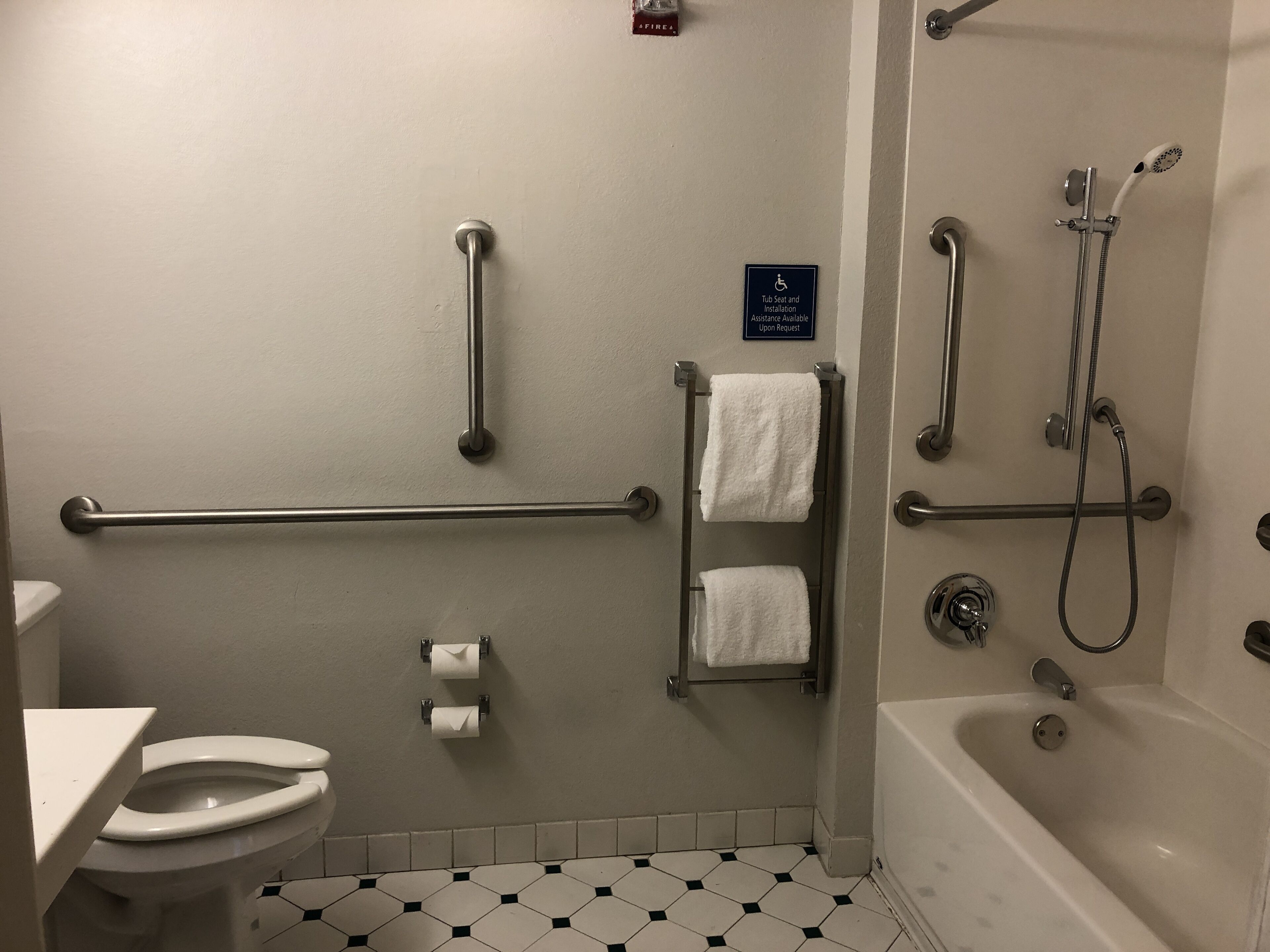 A bathroom with a toilet, handrails, a grab bar, a shower with a handheld showerhead, and a sign indicating assistance is available upon request.