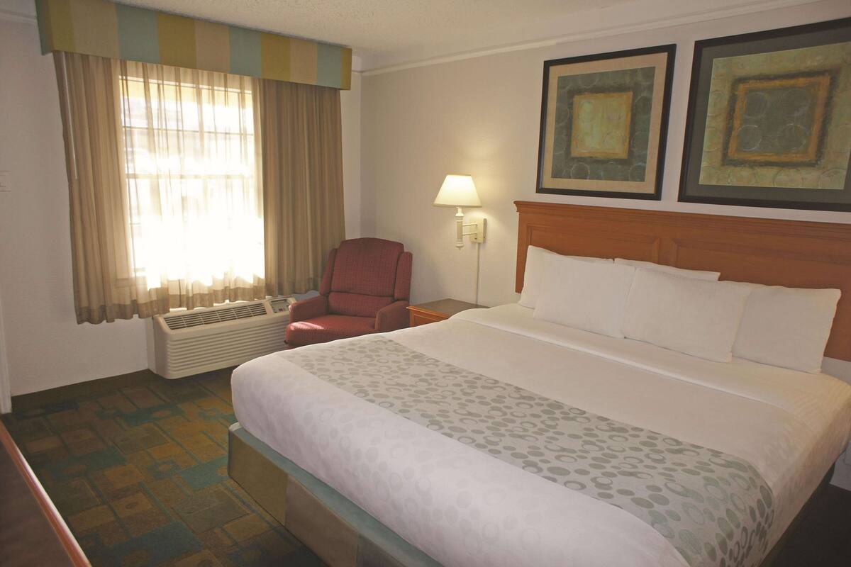 A hotel room with a bed, a chair, a lamp, and a window with curtains.