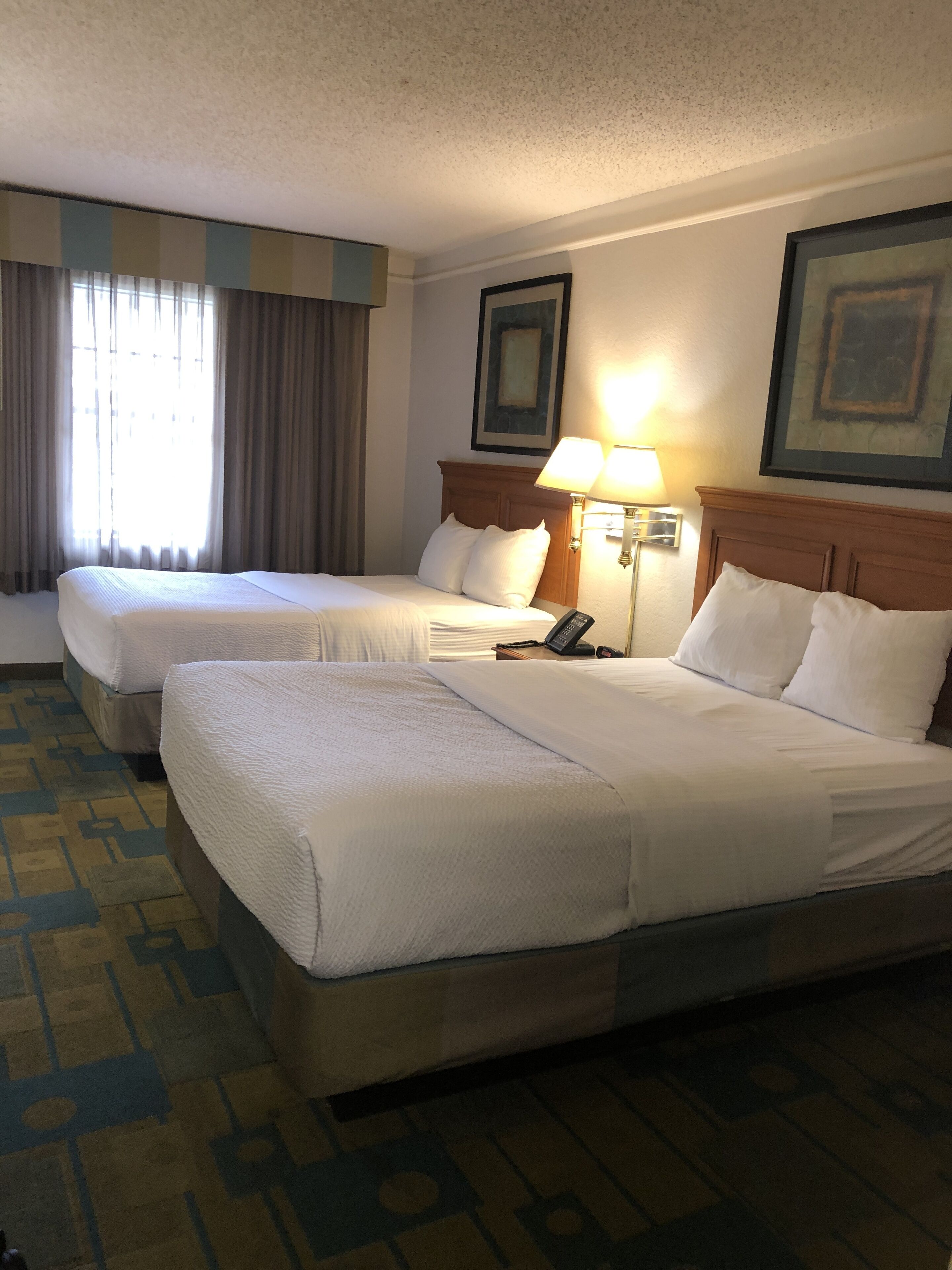 A hotel room with two beds, a nightstand with a lamp, and a window with curtains.