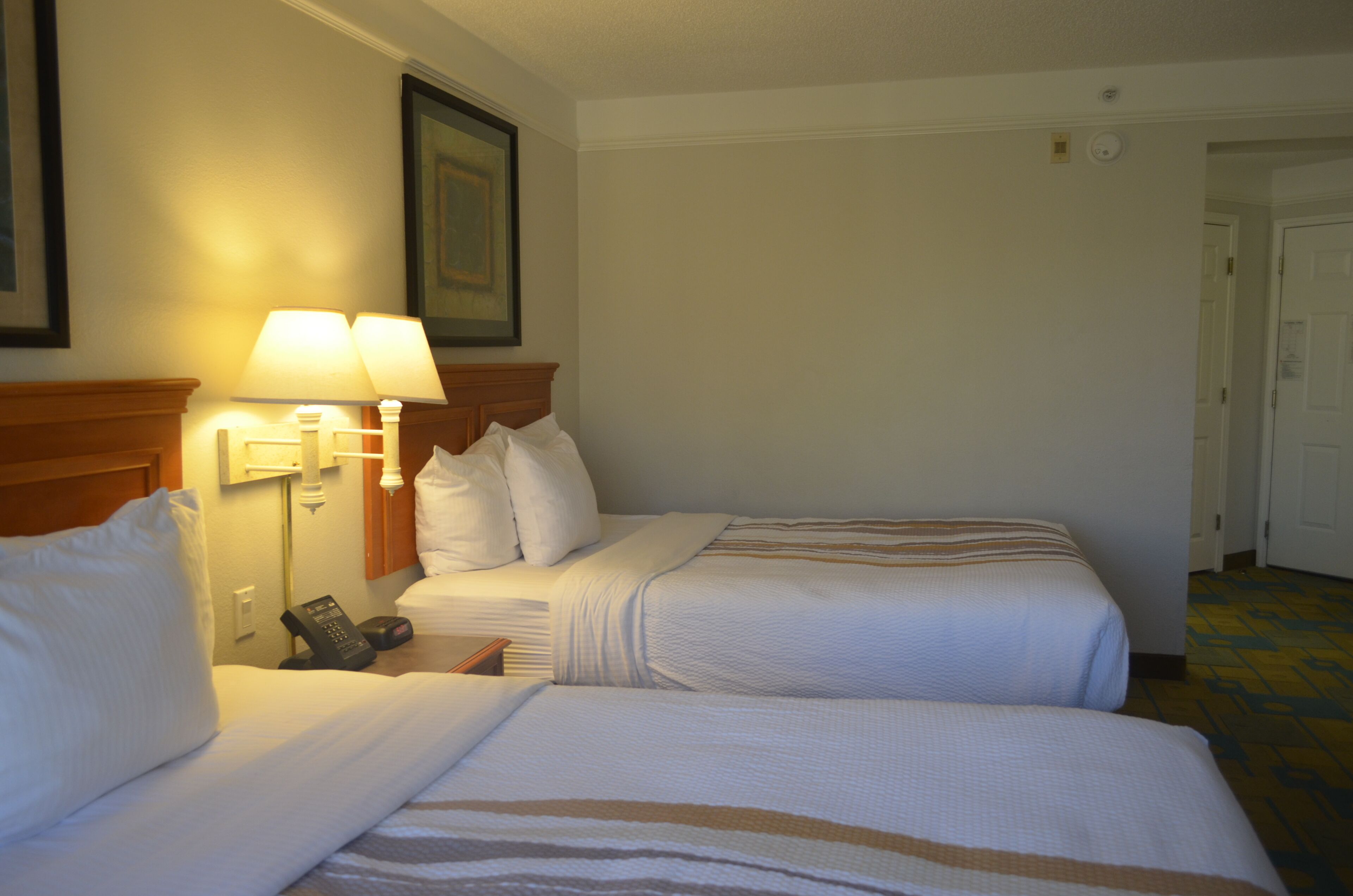 A hotel room with two beds, a nightstand with a lamp, a phone, and a door leading to another room.