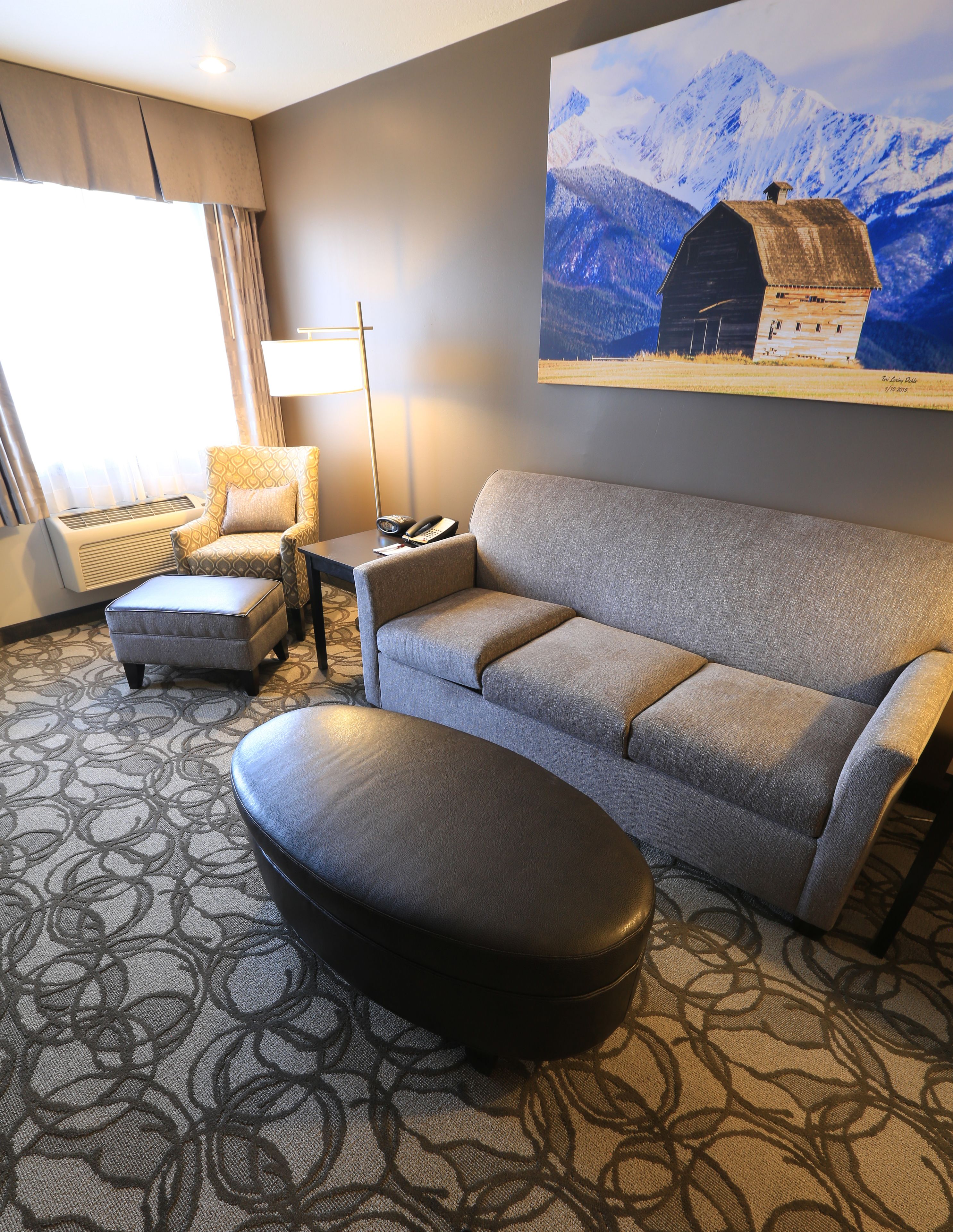 A hotel room with a grey sofa, a matching ottoman, a patterned rug, a standing lamp, a chair, and a painting of a mountain scene.