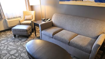 A hotel room with a grey sofa, a matching ottoman, a patterned rug, a standing lamp, a chair, and a painting of a mountain scene.