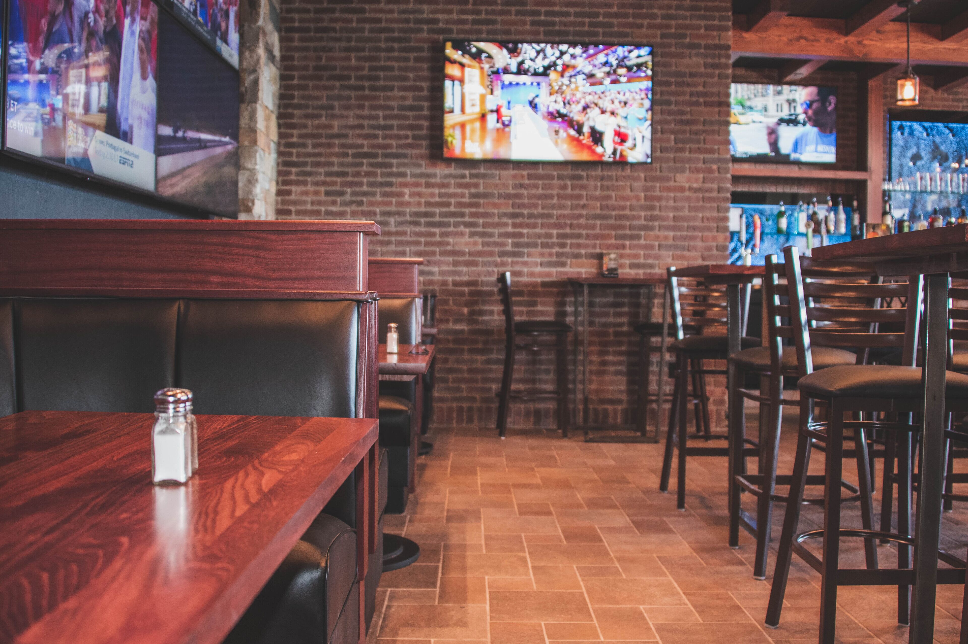 A restaurant interior with brick walls, wooden tables and chairs, and multiple televisions mounted on the wall.
