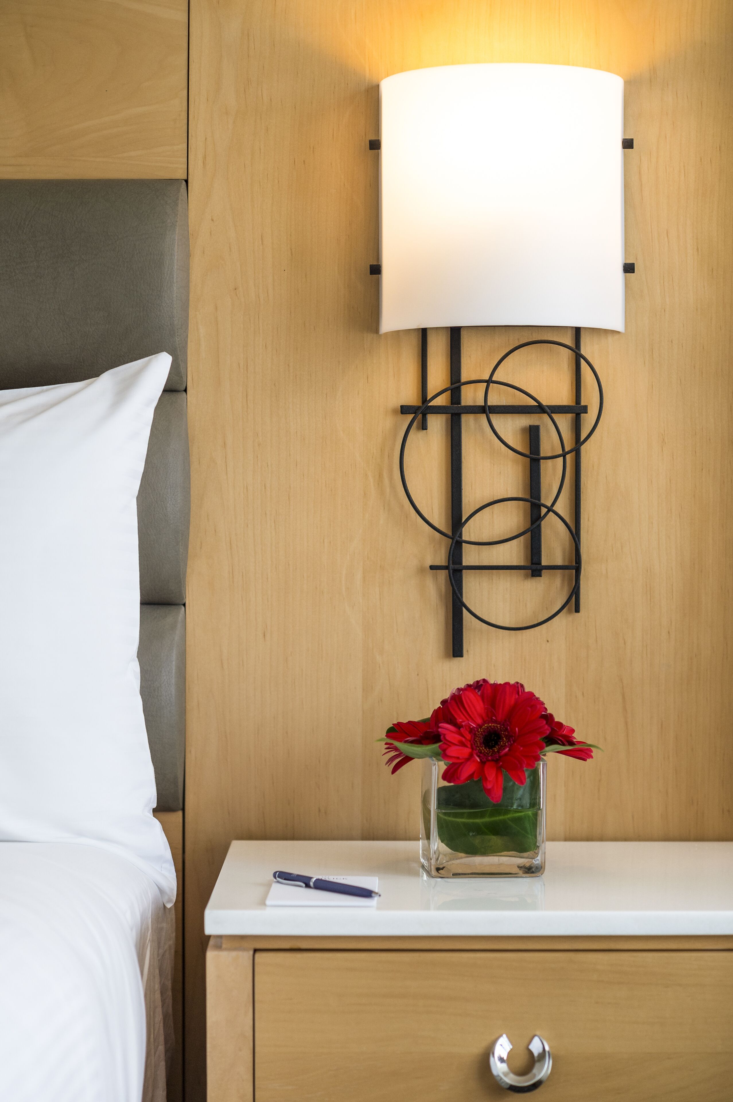 A neatly made bed with white linens, a bedside table with a pen, and a vase with red flowers.