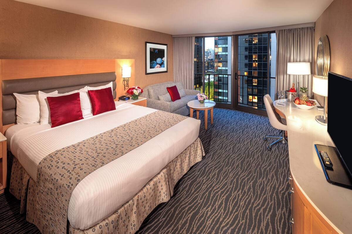 A hotel room with a large bed, a sofa, a small table, a TV, and a view of the city.