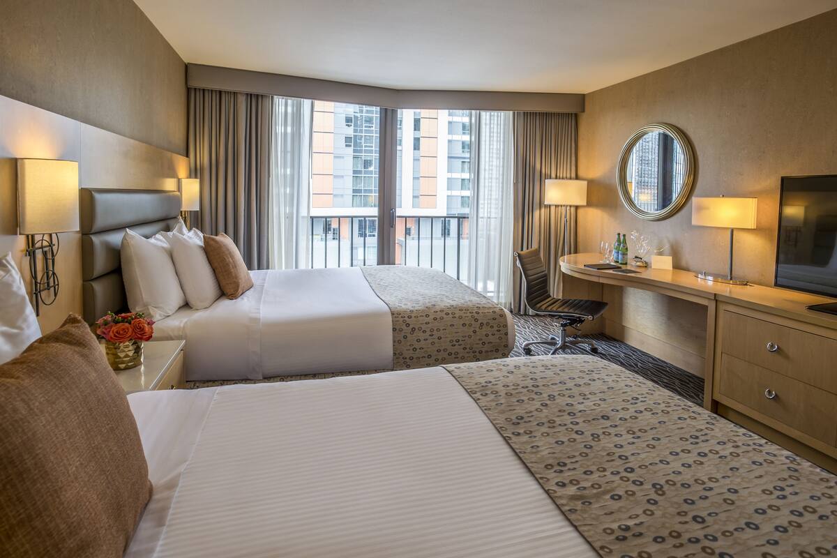 A hotel room with two beds, a desk, a chair, a TV, and a large window with a view of buildings.