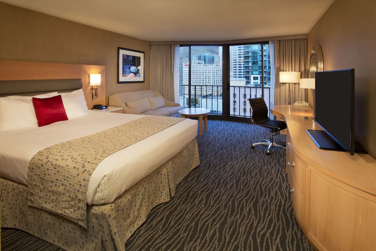 A hotel room with a large bed, a sofa, a desk with a TV, and a view of the city through the windows.