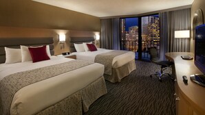 Une chambre d’hôtel avec deux lits, un bureau, une chaise et une vue sur la ville la nuit.