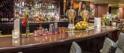 Un espace bar avec un assortiment de bouteilles, une grande tireuse à bière et un panier de fruits. On y trouve des chaises à motifs et un présentoir à menus.
