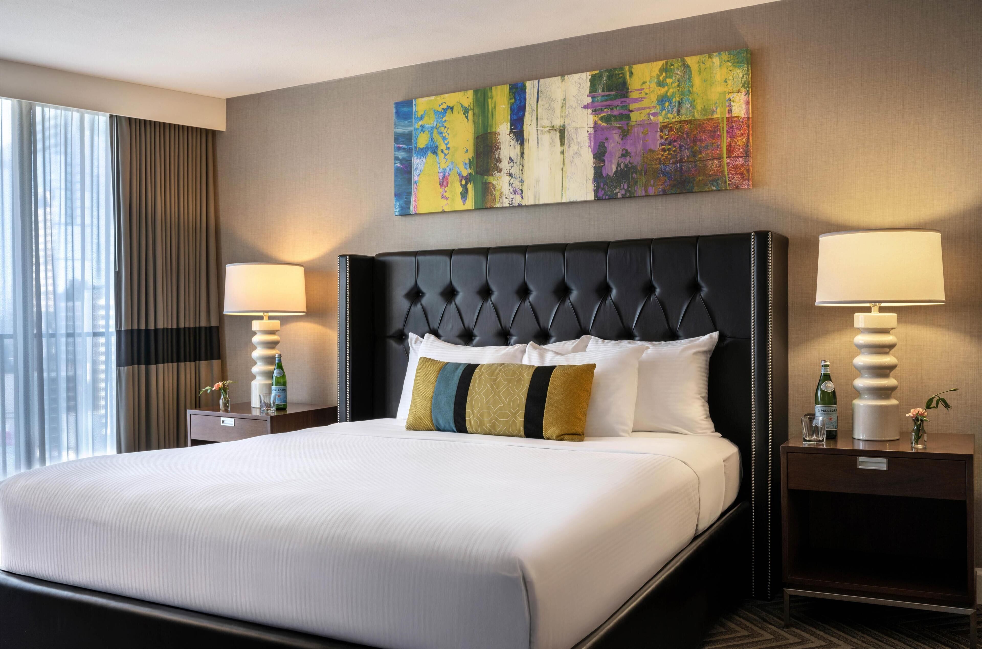 A hotel room with a large bed, two bedside tables, a city view, and a colorful abstract painting above the bed.