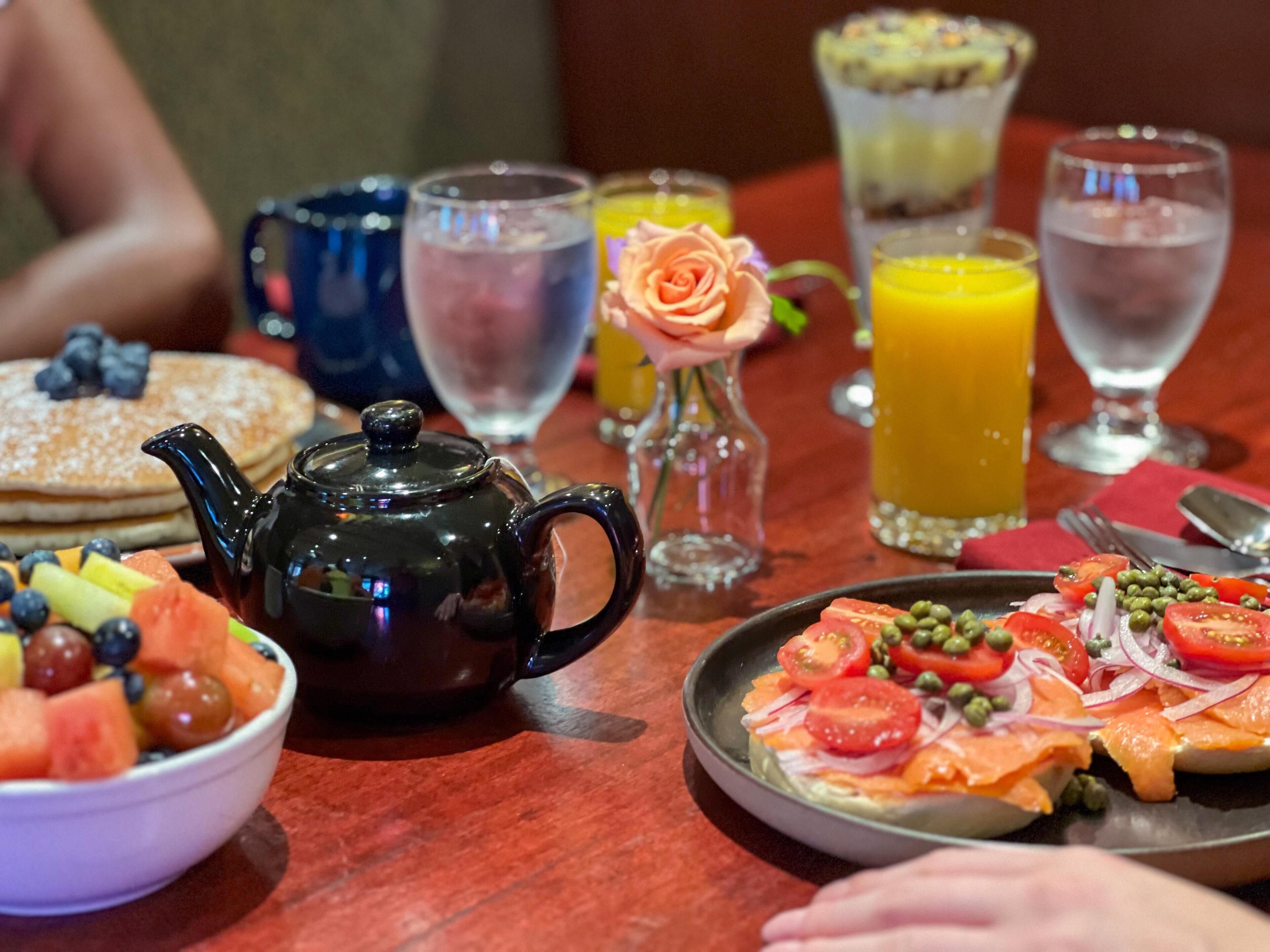 Une table dressée pour le petit-déjeuner avec des crêpes, une théière, des fruits, du jus et une assiette de saumon fumé accompagnée de légumes.