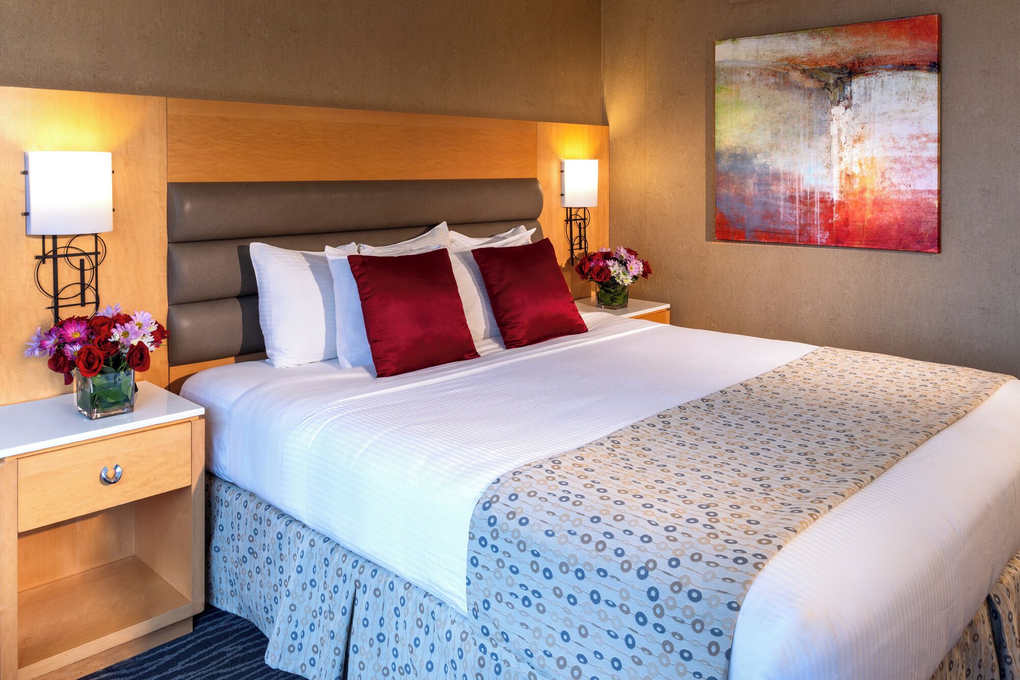 A neatly made bed with a patterned bedspread, two bedside tables with lamps, a vase of flowers, and a painting on the wall.