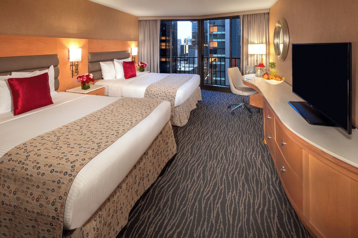A hotel room with two beds, a desk, a TV, and a view of the city.