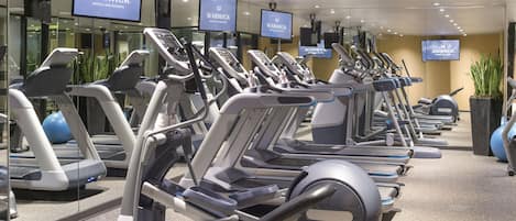 Et fitnesscenter med flere løbebånd og træningsmaskiner, et stort skærm der viser 'Warwick Hotels and Resorts', og en potteplante.
