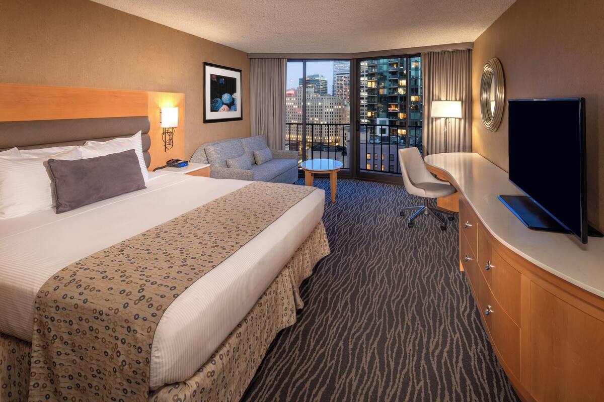 A hotel room with a large bed, a desk, a chair, a TV, and a view of the city through the window.