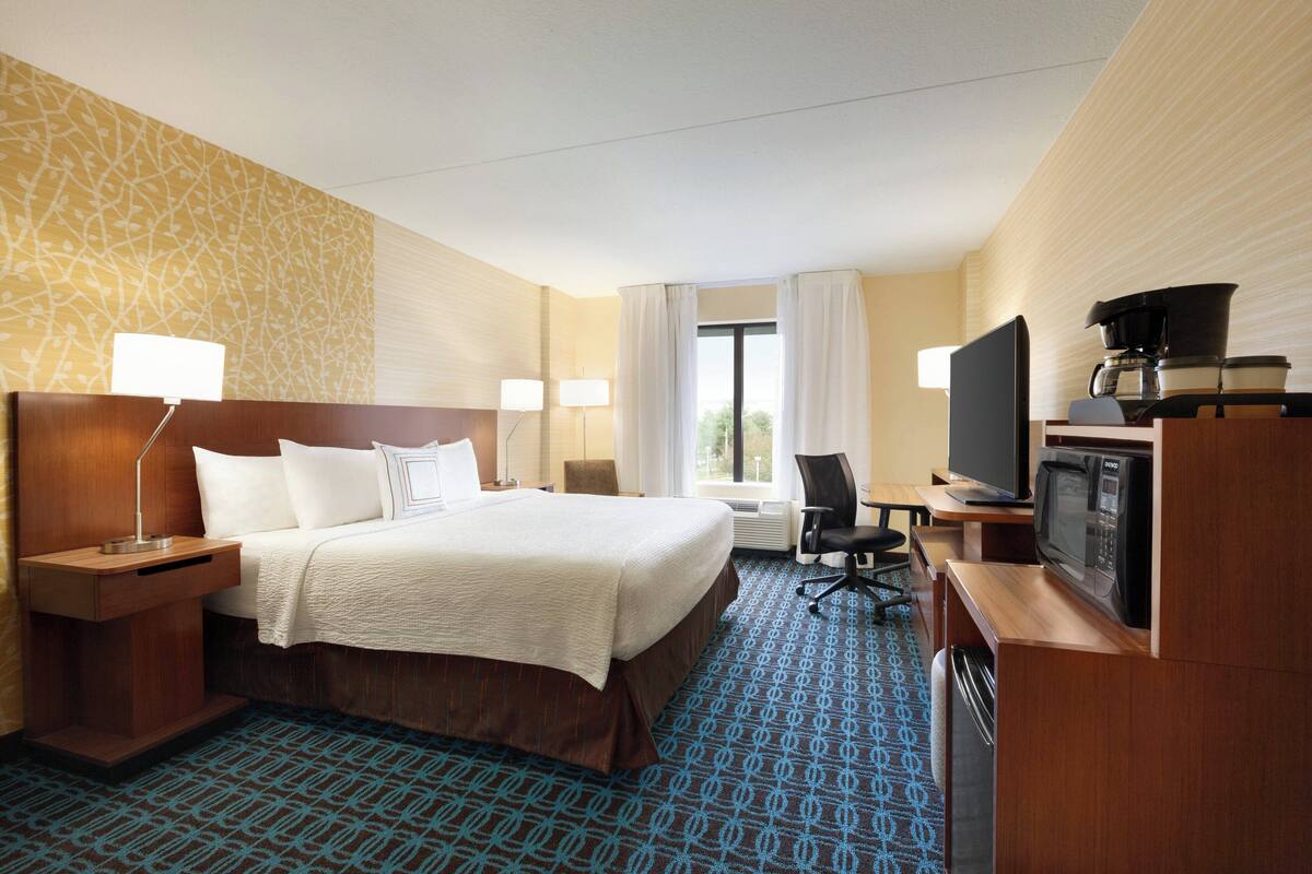 Ein Hotelzimmer mit einem großen Bett, Nachttischen, einem Schreibtisch mit Fernseher und Mikrowelle sowie einem Fenster mit Vorhängen.
