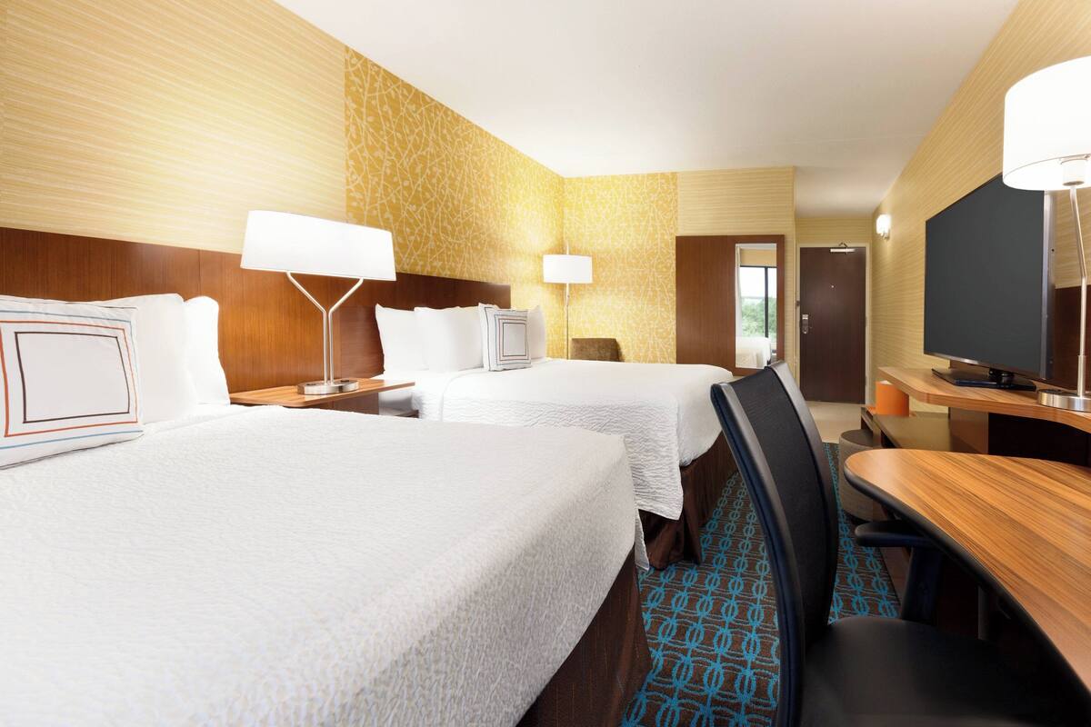 Ein Hotelzimmer mit zwei Betten, einem Schreibtisch, einem Stuhl, einem Fernseher und einer Tür zu einem Badezimmer.