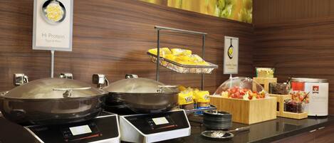 A buffet setup with two hot plates, a fruit display, and a sign indicating 'Hi Protein' with eggs and more.
