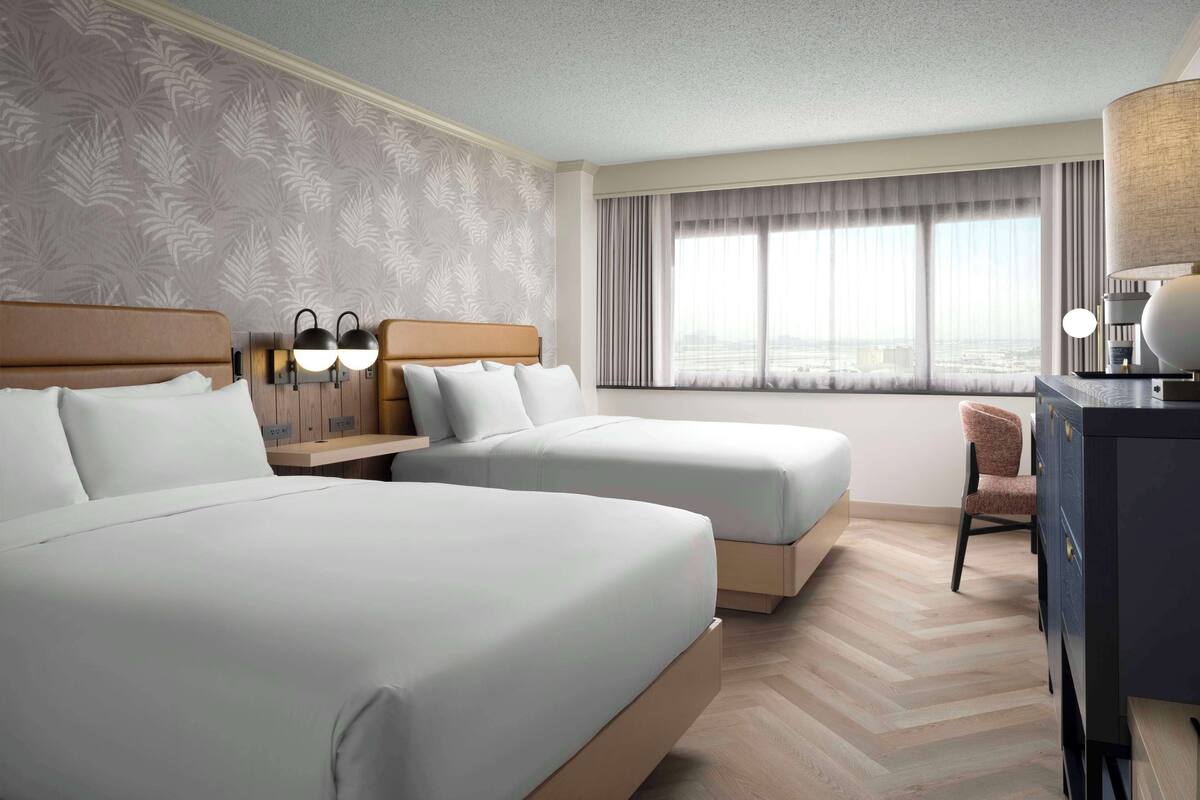 A hotel room with two beds, a desk with a lamp, a chair, and a large window with curtains.