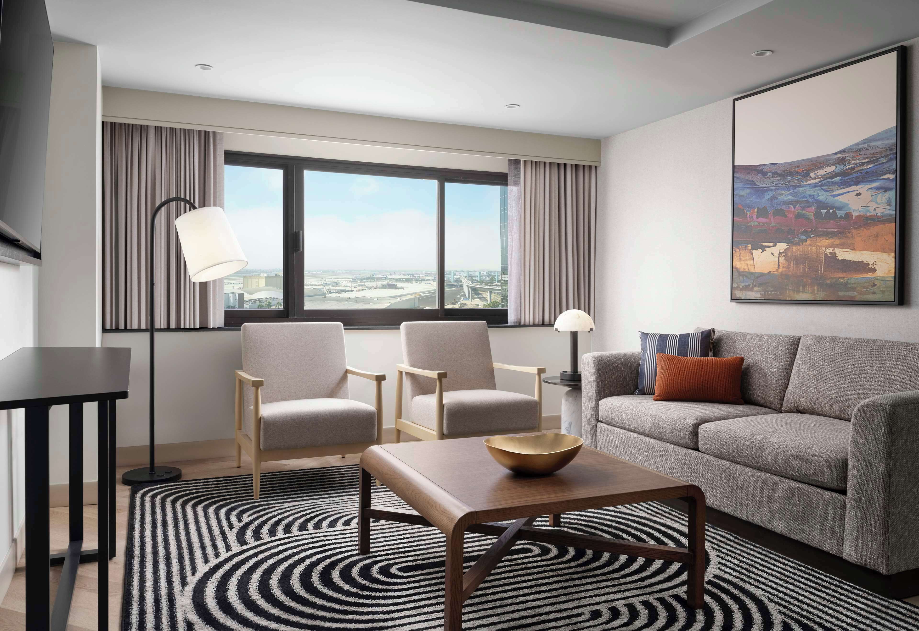 A modern hotel room with a large window, a grey sofa, two armchairs, a coffee table, and a floor lamp.