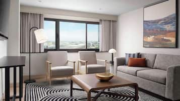 A modern hotel room with a large window, a grey sofa, two armchairs, a coffee table, and a floor lamp.