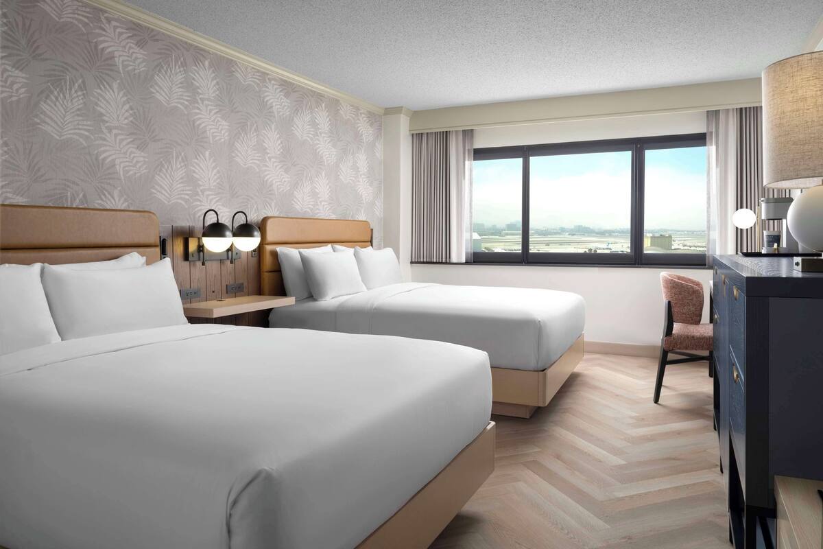 A hotel room with two beds, a desk, a chair, and a large window offering a city view.