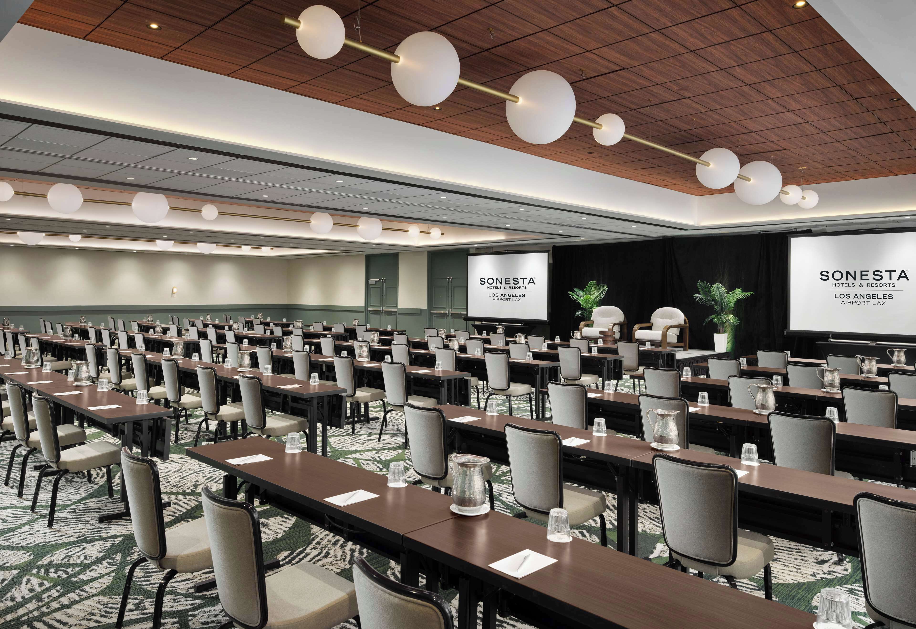 Un salón de conferencias con filas de mesas y sillas, techo de madera con luces colgantes, y una pantalla de proyección que muestra el nombre del hotel.
