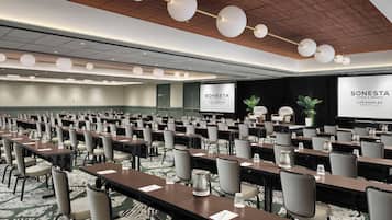 Un salón de conferencias con filas de mesas y sillas, techo de madera con luces colgantes, y una pantalla de proyección que muestra el nombre del hotel.