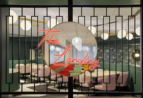 A restaurant interior with a mirrored wall reflecting the room, a neon sign reading "The Landing," and a seating area with round tables and chairs.