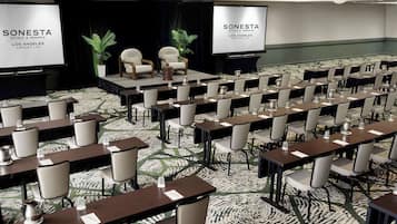 Un salón de conferencias con varias mesas y sillas, un escenario con una pantalla y una alfombra estampada.