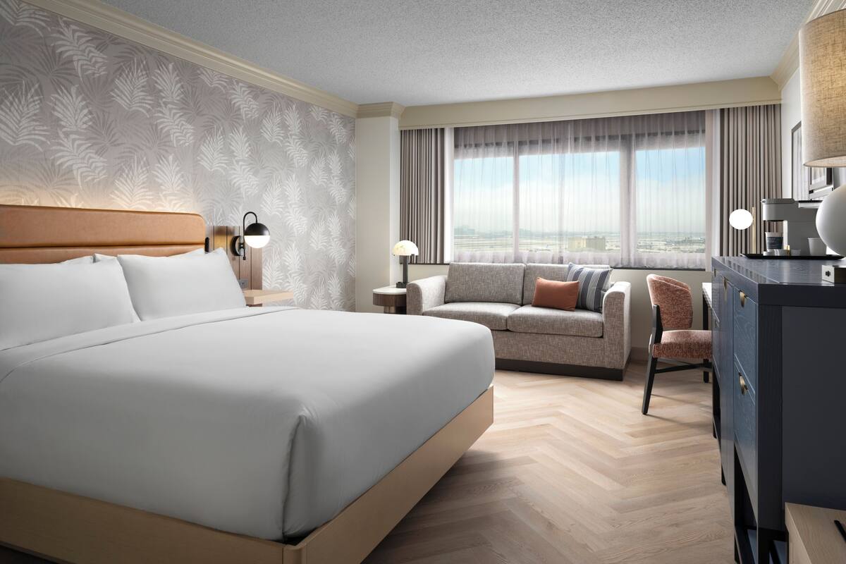 A hotel room with a large bed, a sofa, a chair, a desk, and a view of the city through the window.