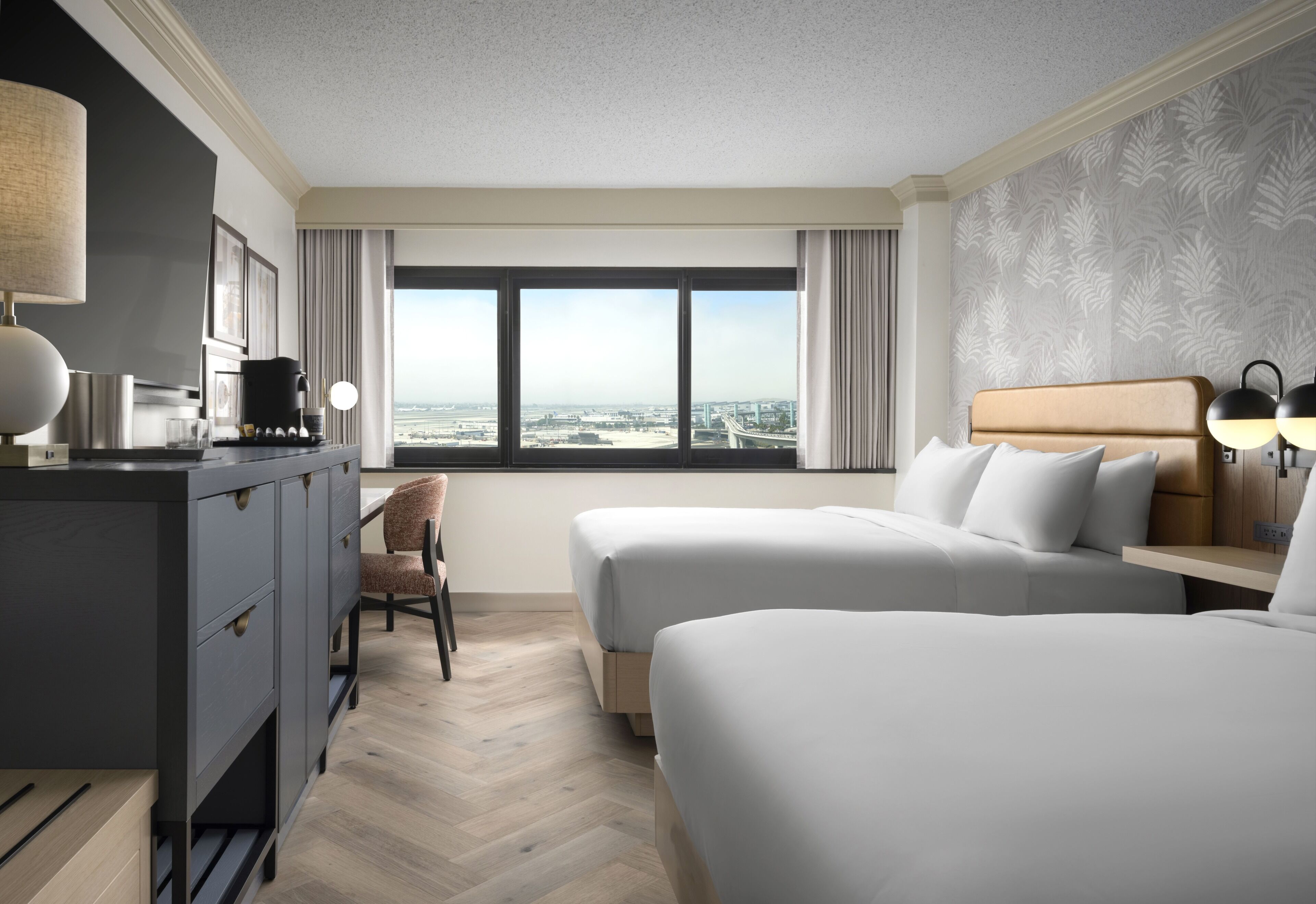 A hotel room with two beds, a desk, a chair, a coffee maker, and a view of the cityscape.