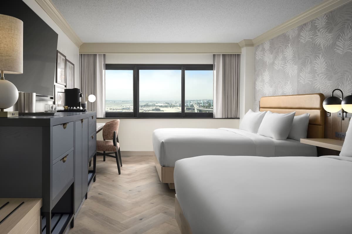 A hotel room with two beds, a desk, a chair, a coffee maker, and a view of the cityscape.