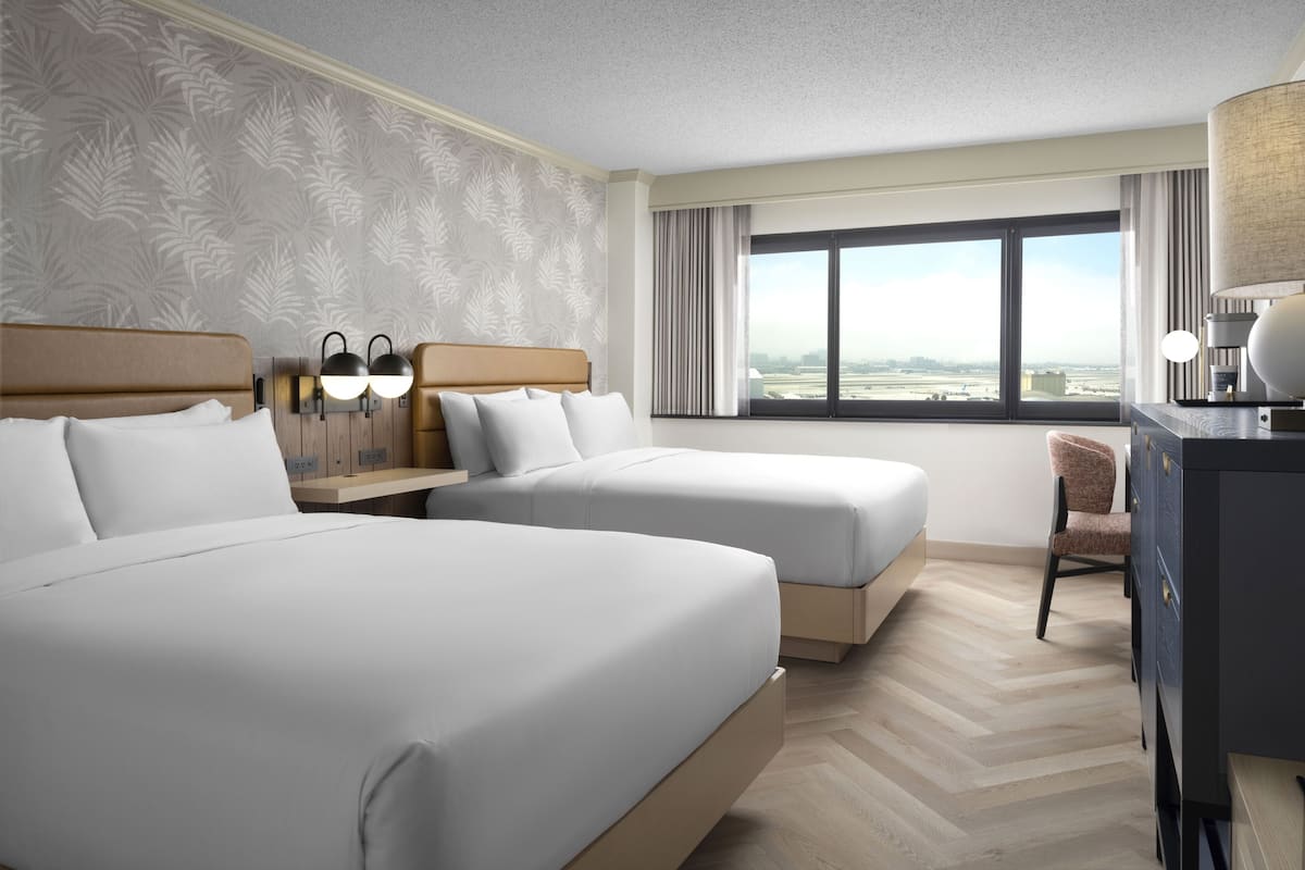 A hotel room with two beds, a desk, a chair, and a large window with a view.