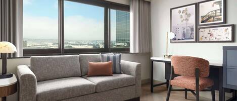 Habitación de hotel moderna con sofá gris, una silla, un escritorio y un ventanal con vistas a la ciudad.