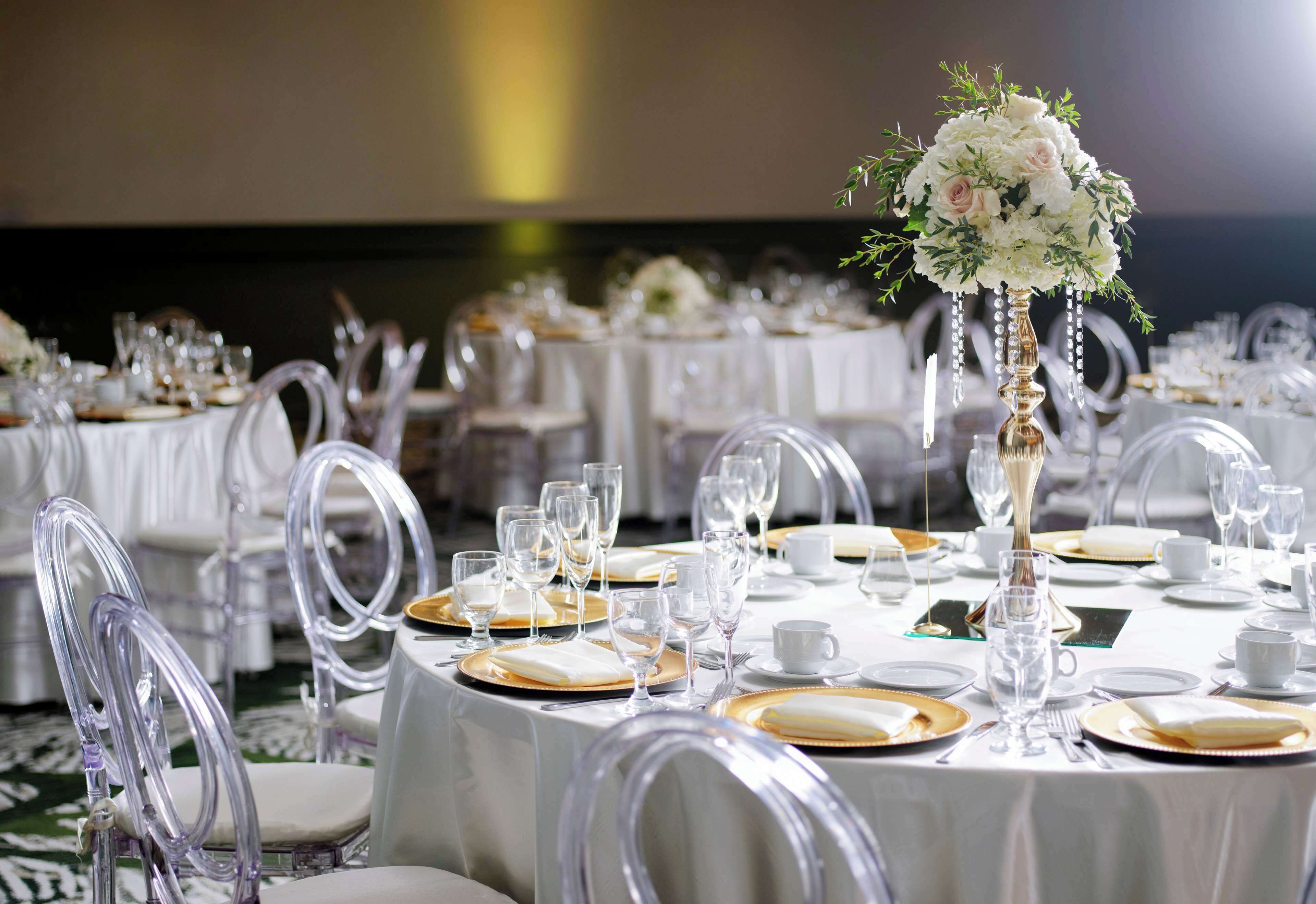 Un salón de banquetes con mesas redondas dispuestas para un evento formal, con sillas transparentes y manteles blancos.