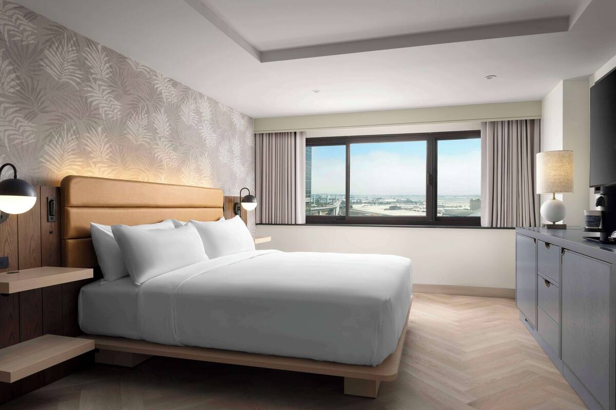 A modern hotel room with a large bed, a nightstand, a desk, a TV, and a window with a city view.