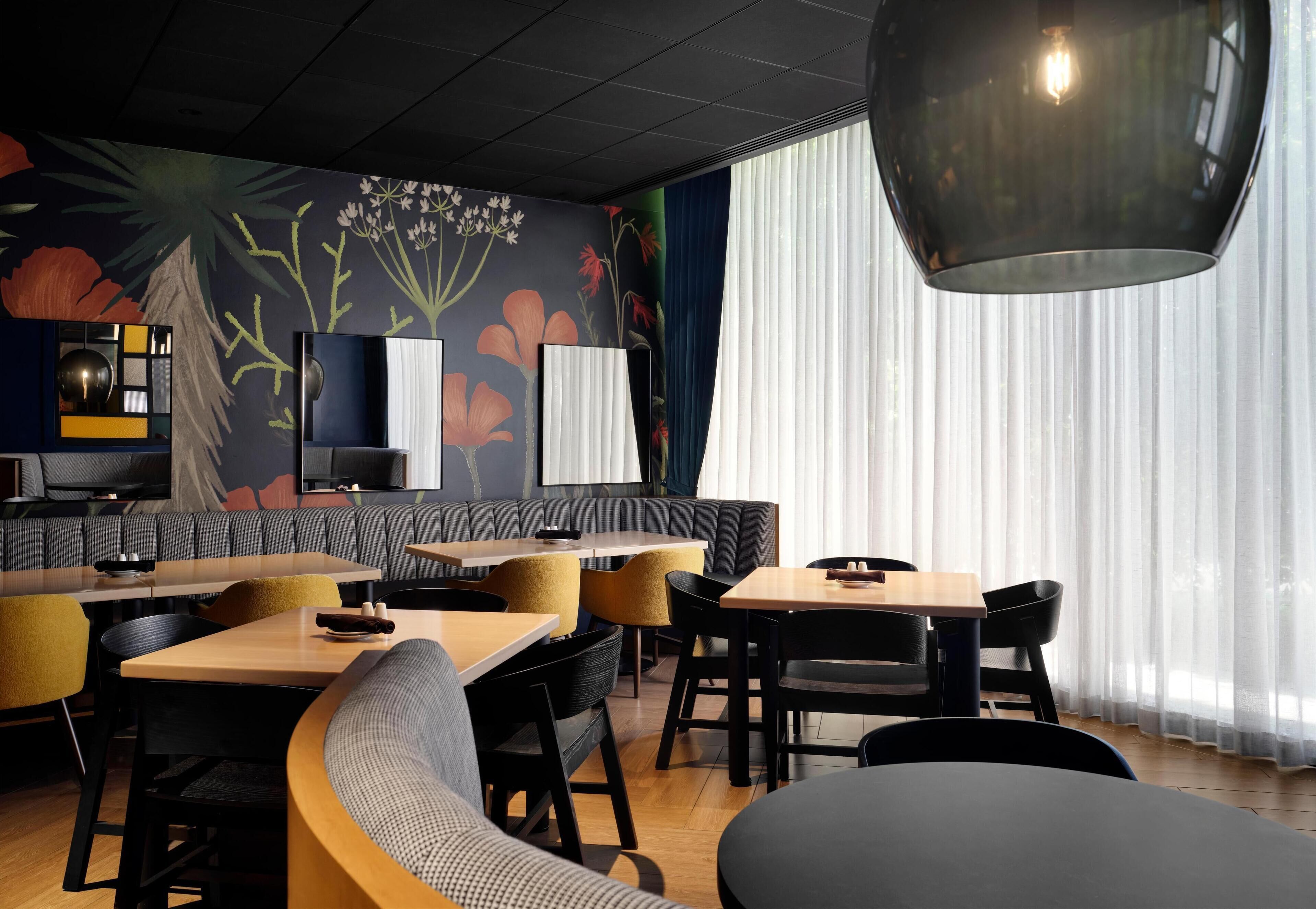 A modern restaurant interior with wooden tables and chairs, a large mural on the wall, and a large pendant light.