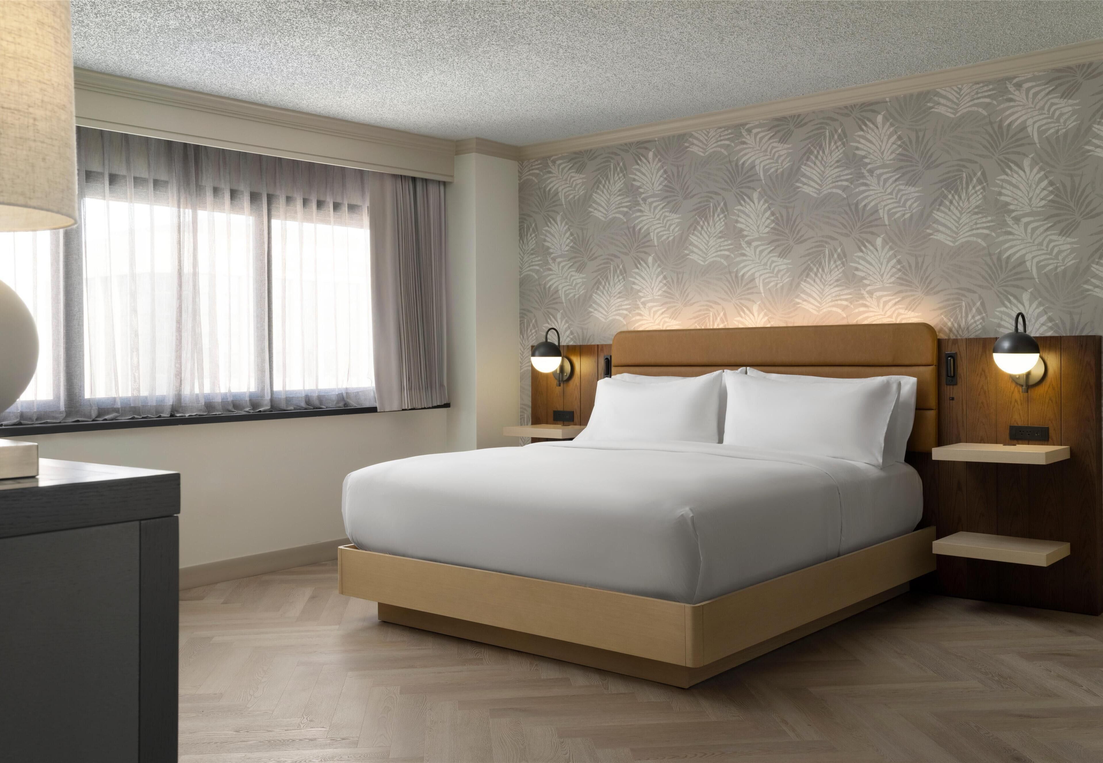 Une chambre d’hôtel moderne avec un grand lit, une tête de lit en bois, deux tables de chevet avec des lampes, une fenêtre avec des rideaux et un papier peint à motifs.