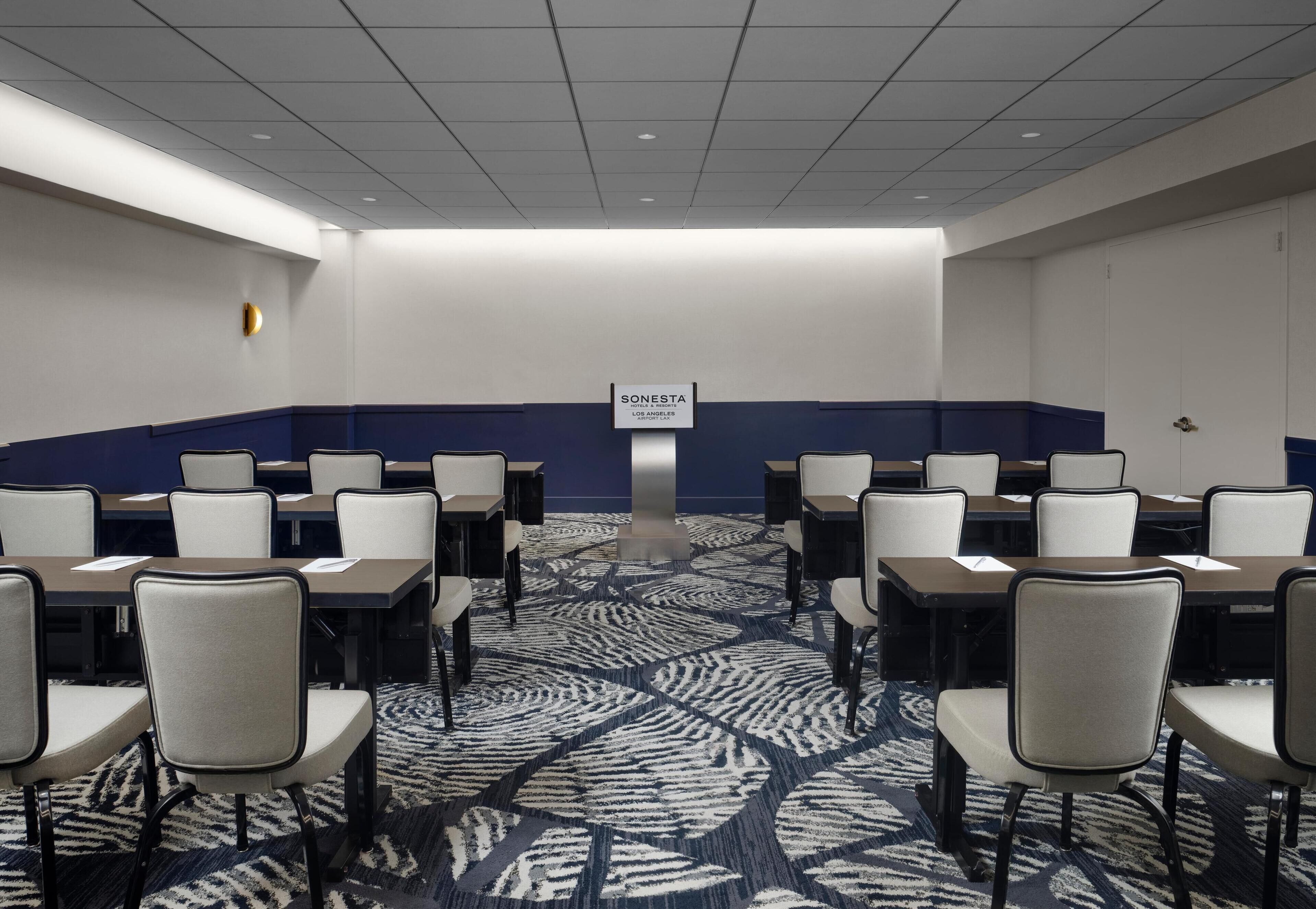A conference room with rows of tables and chairs, a patterned carpet, and a sign indicating it's part of the Sonesta hotel in Los Angeles Airport.