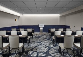 Een vergaderruimte met rijen tafels en stoelen, een kamerbreed tapijt met een patroon, en een bordje dat aangeeft dat het deel uitmaakt van het Sonesta hotel bij Los Angeles Airport.