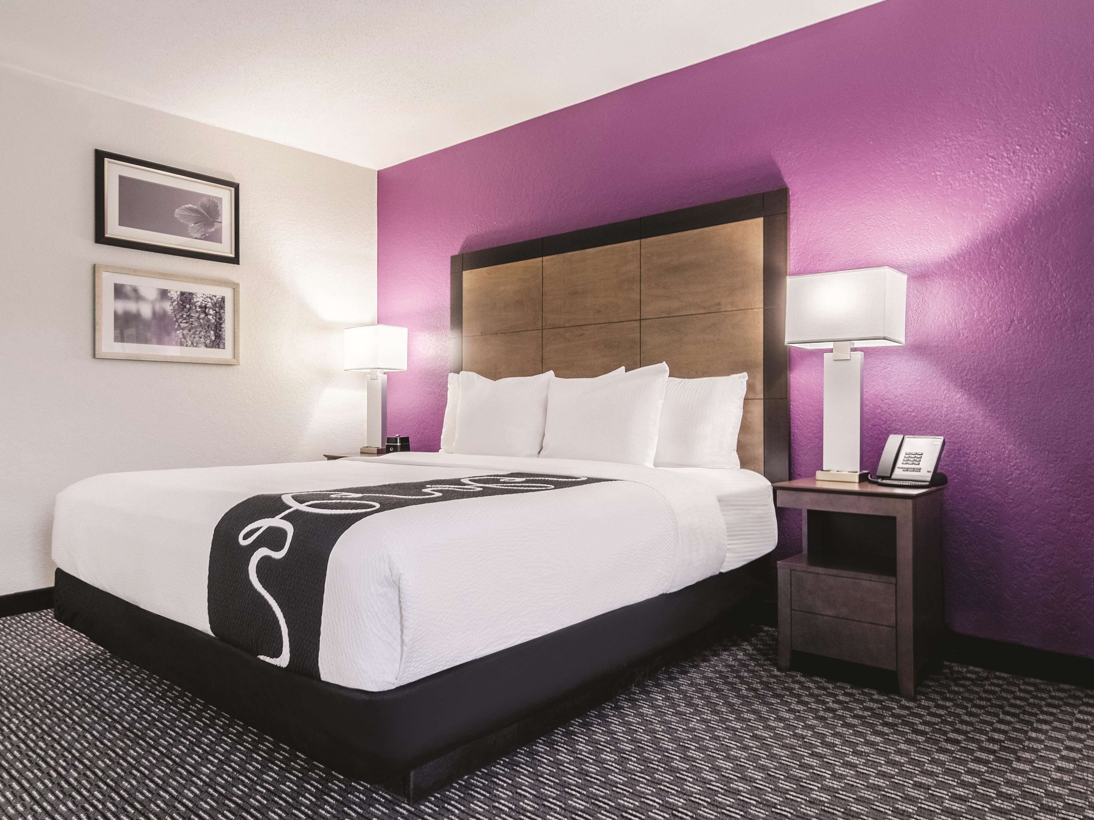 A hotel room with a large bed, two bedside lamps, a nightstand with a phone, and a headboard with a wooden panel.