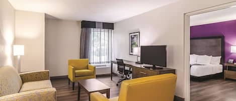 A hotel room with a sofa, two armchairs, a desk with a TV, a bed, and a bathroom visible through an open door.