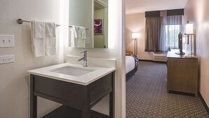 A hotel bathroom with a sink, mirror, towel rack, and a bed visible in the adjacent room.