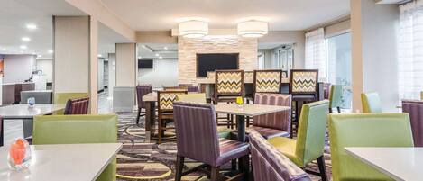 Un espace de restauration moderne dans un hôtel, avec de nombreuses tables et des banquettes, une moquette à motifs et une cheminée centrale.