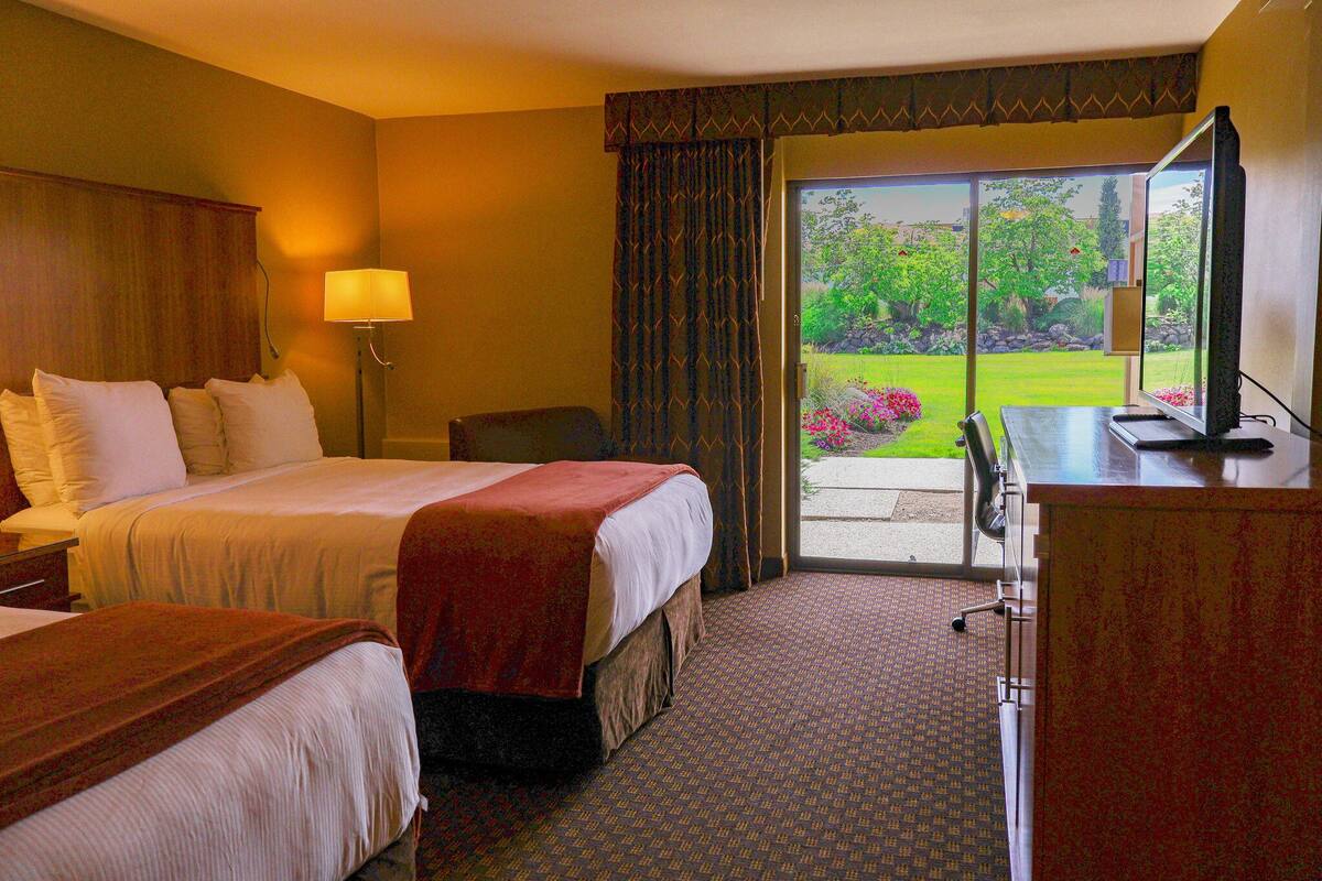 A hotel room with two beds, a desk, a lamp, and a view of a garden through a sliding door.