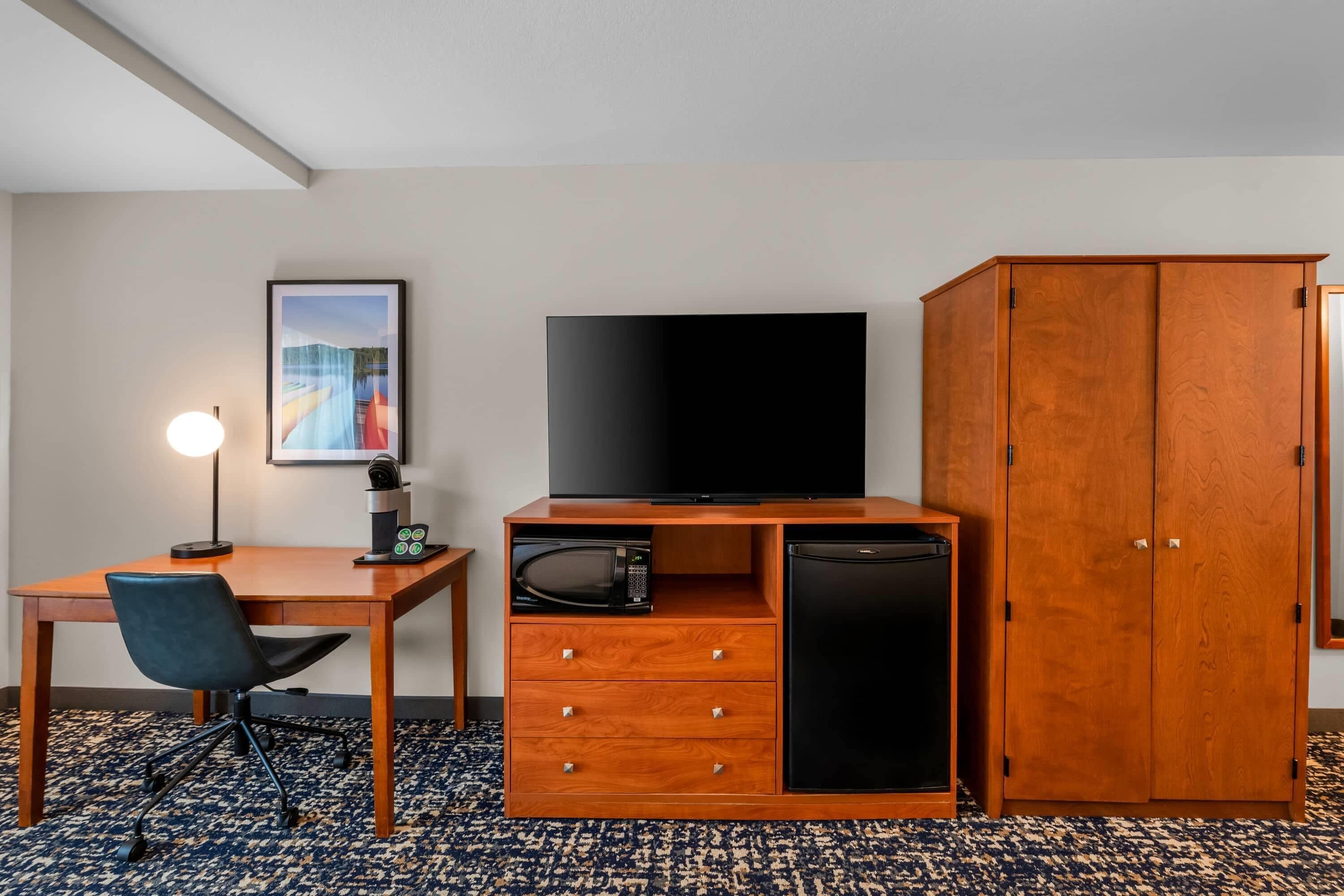 Habitación de hotel con un escritorio de madera, televisión, microondas, refrigerador, una silla y un armario.