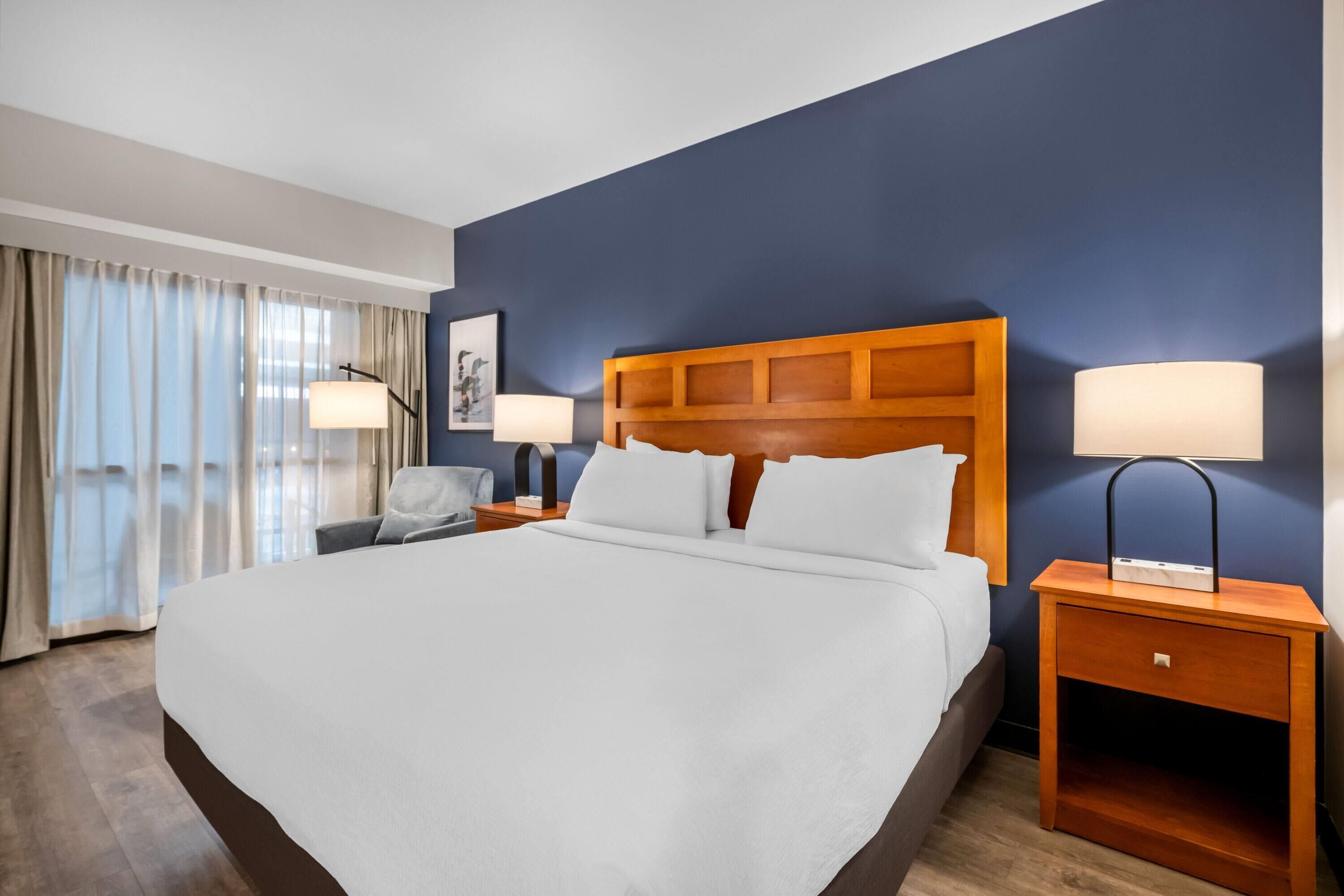 Habitación de hotel con una cama grande, dos mesitas de noche con lámparas, una silla y vistas al exterior.