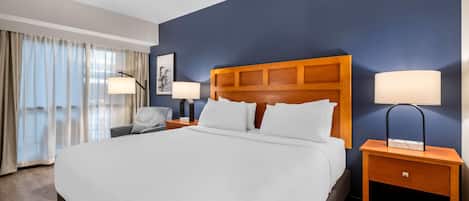 Habitación de hotel con una cama grande, dos mesitas de noche con lámparas, una silla y vistas al exterior.